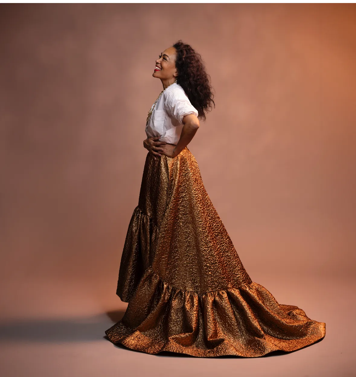 Caroline Henderson står vänd på sidan med händerna på höften framför en ljusbrun bakgrund