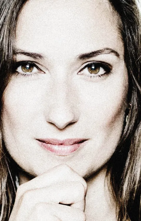 En person med axellångt hår står framför en ljus bakgrund. Personen bär en mörk tröja och håller ena handen upp mot hakan.