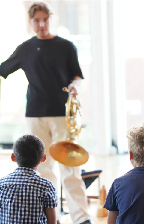 En person håller ett musikinstrument och visar en bild för en grupp barn som sitter framför och lyssnar under en aktivitet inomhus.