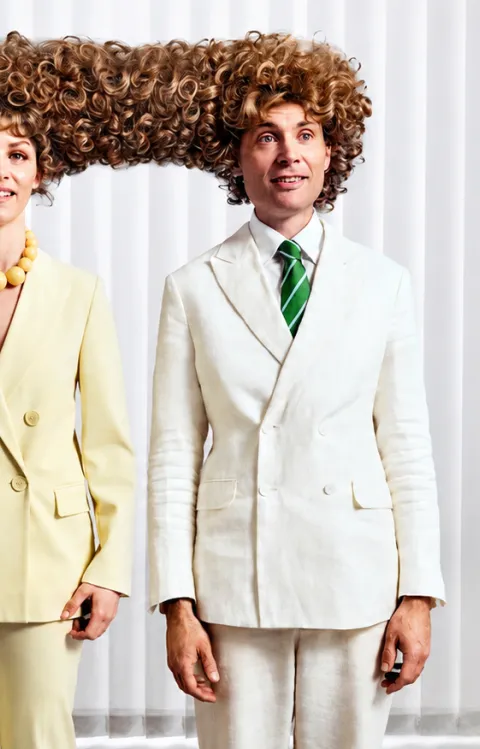 Jesper Rönndahl och Marie Agerhäll står framför vita lamellgardiner i ljusa kostymer, med stort lockigt hår som humoristiskt binder samman deras frisyrer.