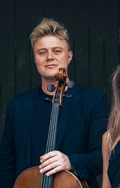 Tre musiker står framför en mörk trävägg med cello och violin, klädda i mörka konsertkläder.