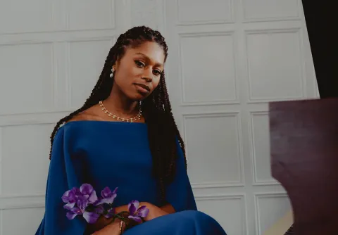 Person i blå klänning sitter vid ett piano och håller lila blommor framför vit panelvägg till konserten Stravinskij och Beethoven med Malmö Symfoniorkester.