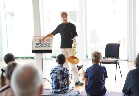 En person håller ett musikinstrument och visar en bild för en grupp barn som sitter framför och lyssnar under en aktivitet inomhus till konserten Eldfågeln med Malmö Symfoniorkester.