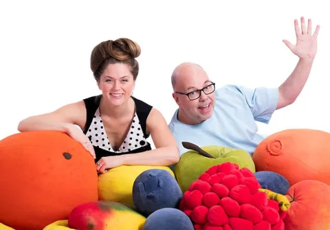 Mimi Terris och Dan Bornemark ler mot kameran bland stora färgglada frukter mot en vit bakgrund.