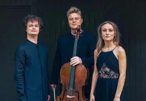 Tre musiker står framför en mörk trävägg med cello och violin, klädda i mörka konsertkläder.